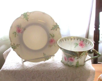 May include: A vintage teacup and saucer set. The cup and saucer are cream-colored with pink rose and green leaf designs. The cup has a gold trim and a green handle. The saucer is supported by a white metal stand.