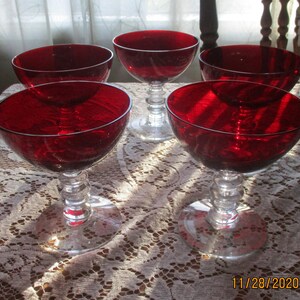 Pu&ograve; includere: Set di sei coppe da champagne in vetro rosso con steli in vetro trasparente. Le coppe sono disposte su un tovagliolo di pizzo bianco.