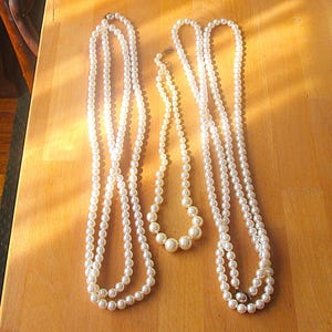 May include: Three strands of faux pearl necklaces of varying lengths rest on a wooden surface. The necklaces are white and have a classic, elegant appearance. The pearls are round and evenly spaced, creating a timeless jewelry aesthetic.