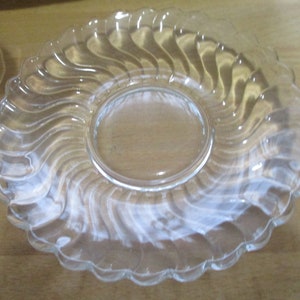 May include: A clear glass saucer with a wavy, swirl design. The saucer has a smooth, flat surface in the center.