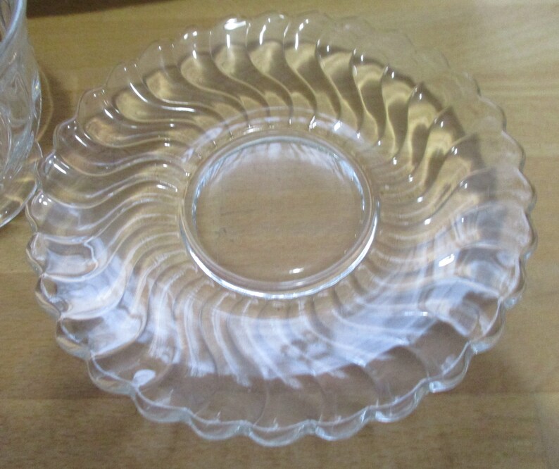 May include: A clear glass saucer with a wavy, swirl design. The saucer has a smooth, flat surface in the center.