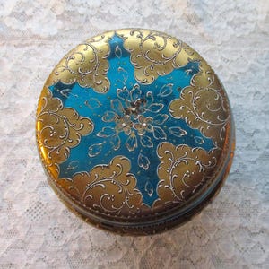 Op de afbeelding: Een ronde, vintage sieradendoos met een goud en blauwgroen ontwerp. Het deksel heeft een blauwgroen midden met een bloemmotief en gouden versieringen in filigraanstijl. De zijkanten van de doos zijn ook goudkleurig.