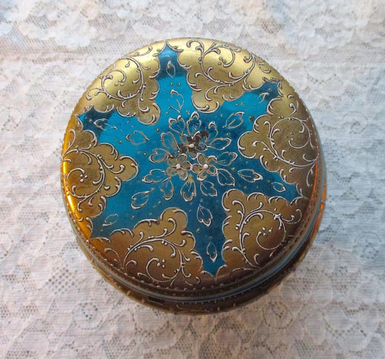 Op de afbeelding: Een ronde, vintage sieradendoos met een goud en blauwgroen ontwerp. Het deksel heeft een blauwgroen midden met een bloemmotief en gouden versieringen in filigraanstijl. De zijkanten van de doos zijn ook goudkleurig.