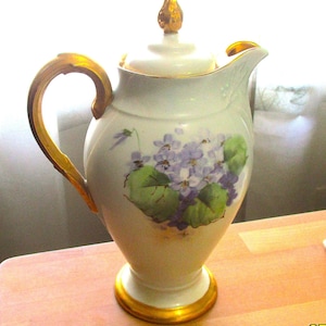 May include: A white porcelain coffee pot with a gold handle, spout, and base. The pot features a floral design of purple violets and green leaves. The lid is topped with a gold finial.