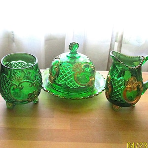 May include: A set of three green glass serving pieces with gold accents. The set includes a butter dish with a lid, a creamer, and a sugar bowl. The pieces are decorated with a diamond pattern and floral motifs.