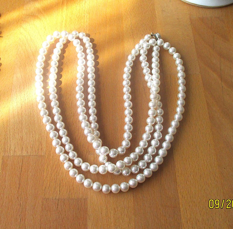 May include: A multi-strand pearl necklace with a silver clasp. The necklace features three strands of round, white pearls, creating a classic and elegant look. The pearls are evenly sized and have a subtle luster, perfect for formal occasions.