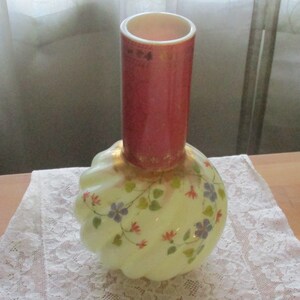 May include: A white glass vase with a bulbous base and a slender neck. The vase is decorated with a floral pattern in shades of red, orange, and purple. The neck of the vase is a solid red color with gold accents.