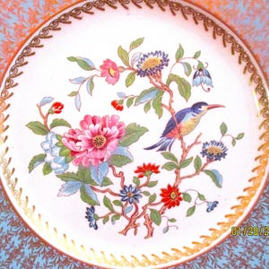 May include: A white porcelain plate with a gold rim and a blue and gold border. The center of the plate features a colorful floral design with a blue bird perched on a branch.