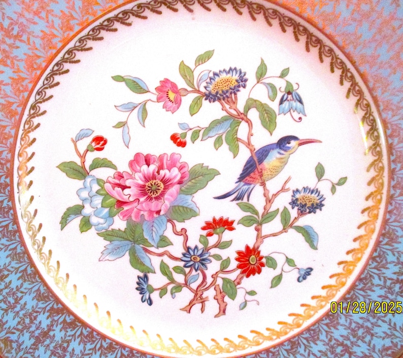 May include: A white porcelain plate with a gold rim and a blue and gold border. The center of the plate features a colorful floral design with a blue bird perched on a branch.