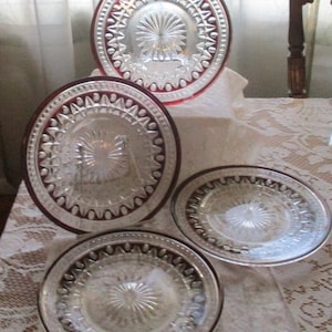 May include: Set of four clear glass plates with a red rim and a starburst pattern in the center. The plates are stacked on top of each other.