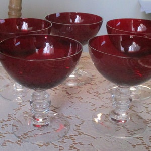 Pu&ograve; includere: Set di sei coppette da dessert in vetro rosso con steli in vetro trasparente. Le coppette sono disposte su un tovagliolo di pizzo bianco.