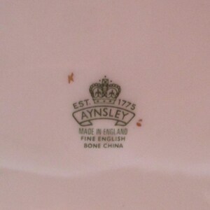 May include: A close-up of a white porcelain plate with a green and gold logo that reads "Est. 1775 Aynsley Made in England Fine English Bone China".
