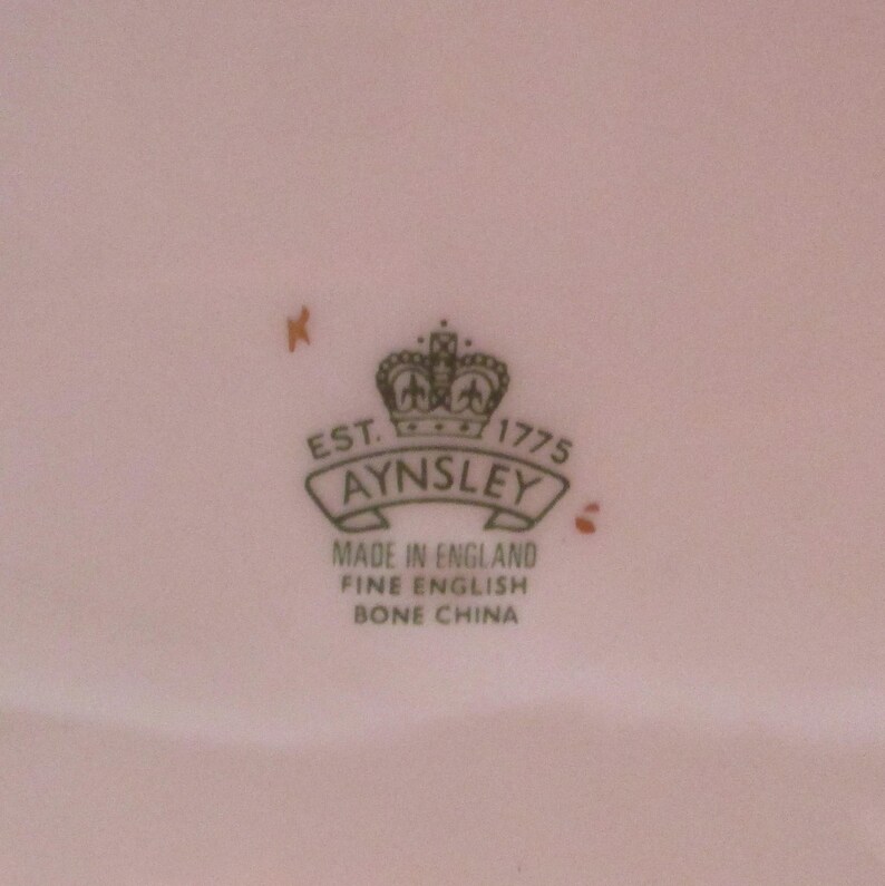 May include: A close-up of a white porcelain plate with a green and gold logo that reads "Est. 1775 Aynsley Made in England Fine English Bone China".