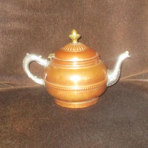 May include: A copper teapot with a silver handle and spout. The lid is copper with a gold finial. The teapot has a rounded body with decorative detailing. The handle and spout have ornate designs.