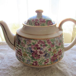 May include: A white teapot with a floral pattern and gold trim. The teapot has a lid and a handle.