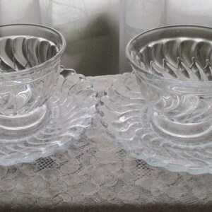 May include: Two clear glass teacups with swirling designs on the inside of the cups. Each cup is on a matching clear glass saucer with a swirling design.