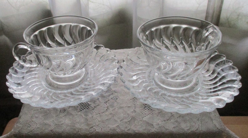 May include: Two clear glass teacups with swirling designs on the inside of the cups. Each cup is on a matching clear glass saucer with a swirling design.