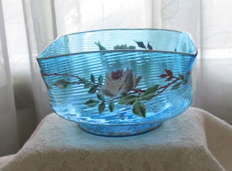 May include: A blue glass bowl with a white rose and green leaves painted on the side. The bowl has a rippled texture.