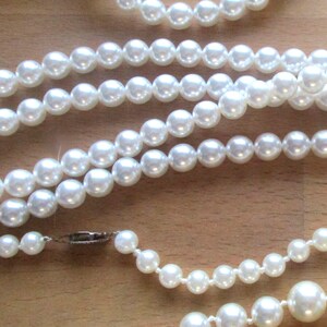 May include: Several strands of white faux pearl necklaces and a bracelet are displayed on a wooden surface. The pearls vary in size, with a clasp visible on one necklace. The jewelry is arranged in a layered fashion.