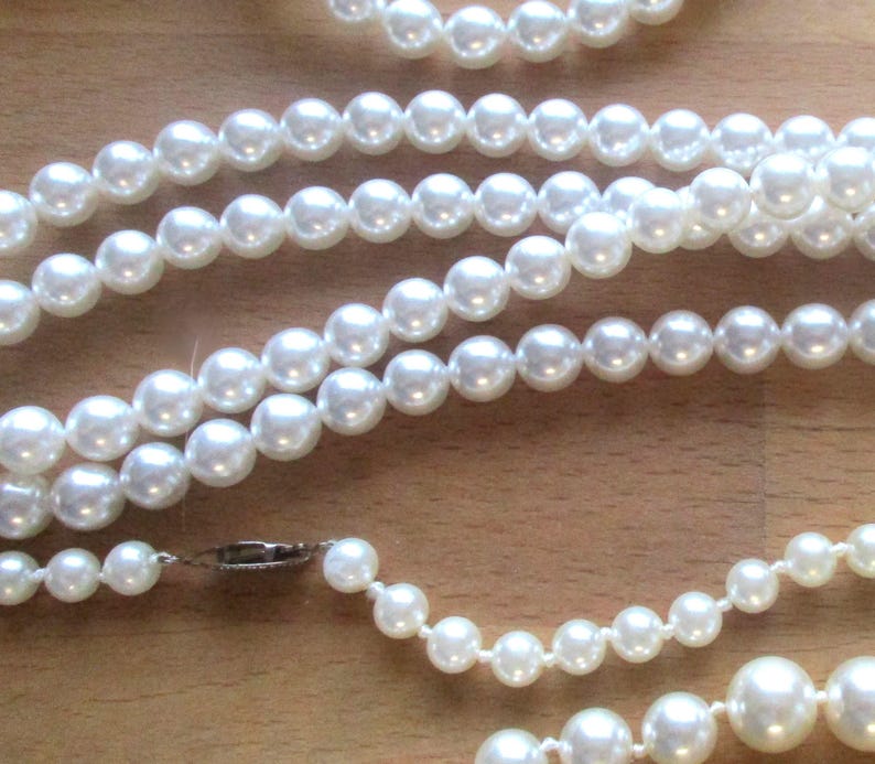May include: Several strands of white faux pearl necklaces and a bracelet are displayed on a wooden surface. The pearls vary in size, with a clasp visible on one necklace. The jewelry is arranged in a layered fashion.