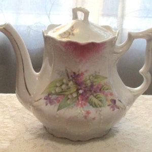 May include: White porcelain teapot with a floral design featuring purple and white flowers. The teapot has a gold trim around the spout and handle.