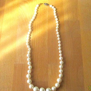 May include: A cream-colored pearl necklace with varying sizes of round beads. The necklace is fastened with a small clasp and lies on a wooden surface. The pearls graduate in size, with the largest at the center.