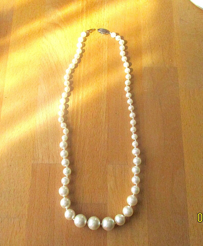 May include: A cream-colored pearl necklace with varying sizes of round beads. The necklace is fastened with a small clasp and lies on a wooden surface. The pearls graduate in size, with the largest at the center.