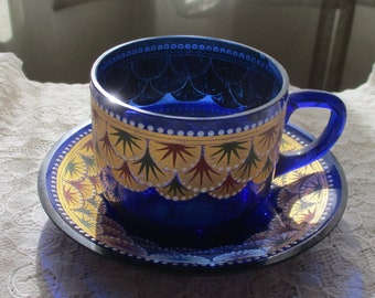 May include: A blue glass teacup and saucer with a gold and red geometric pattern. The cup and saucer are decorated with a repeating design of stylized leaves and dots.