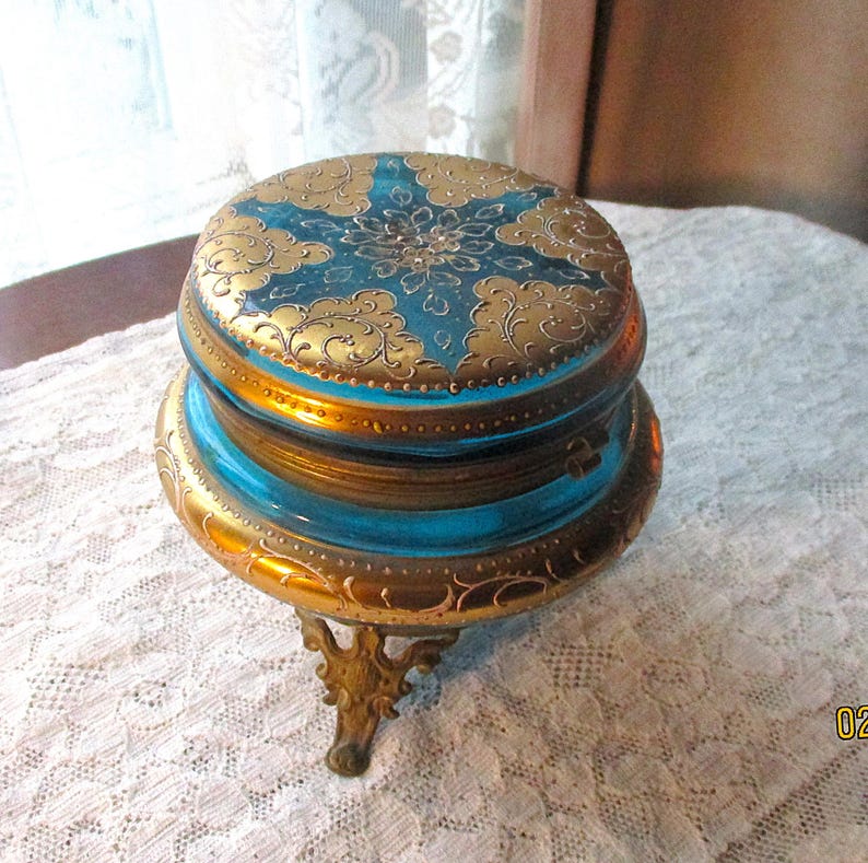 Op de afbeelding: Een antieke, sierlijke sieradendoos met een teal en goud kleurenschema. Het deksel en de basis hebben ingewikkelde gouden filigraan ontwerpen op een teal achtergrond. De doos staat op een decoratieve, driepootstandaard, wat bijdraagt aan de vintage charme.