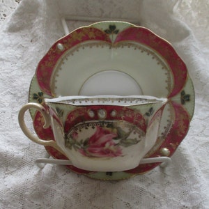 May include: A vintage teacup and saucer set with a floral design. The cup and saucer are decorated with pink roses, green leaves, and gold accents. The saucer has a scalloped edge.