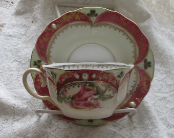 May include: A vintage teacup and saucer set with a floral design. The cup and saucer are decorated with pink roses, green leaves, and gold accents. The saucer has a scalloped edge.