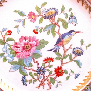 May include: A white porcelain plate with a gold rim and a floral design featuring pink, red, blue, and green flowers and a blue and yellow bird.