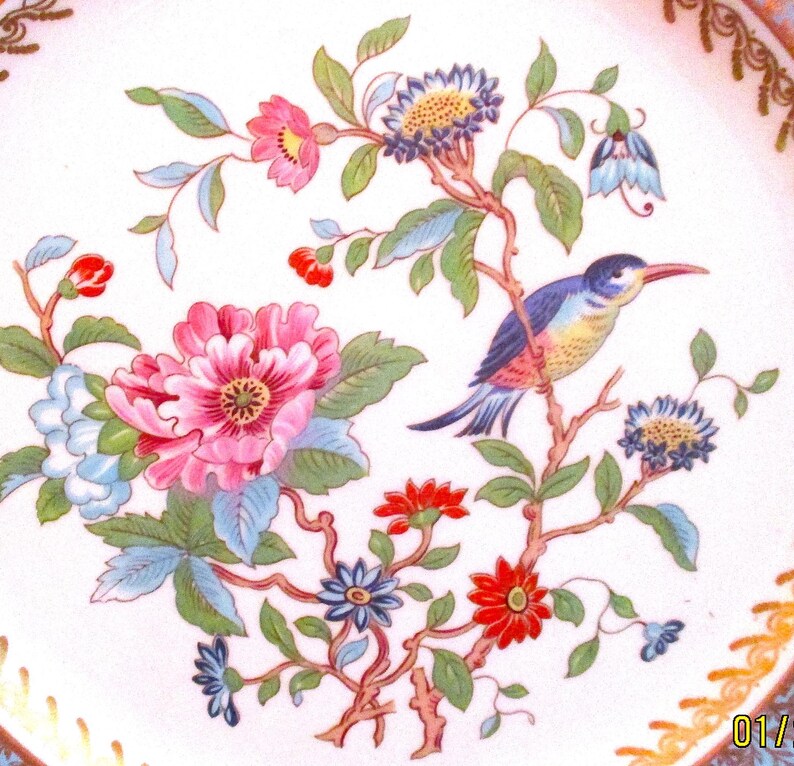 May include: A white porcelain plate with a gold rim and a floral design featuring pink, red, blue, and green flowers and a blue and yellow bird.