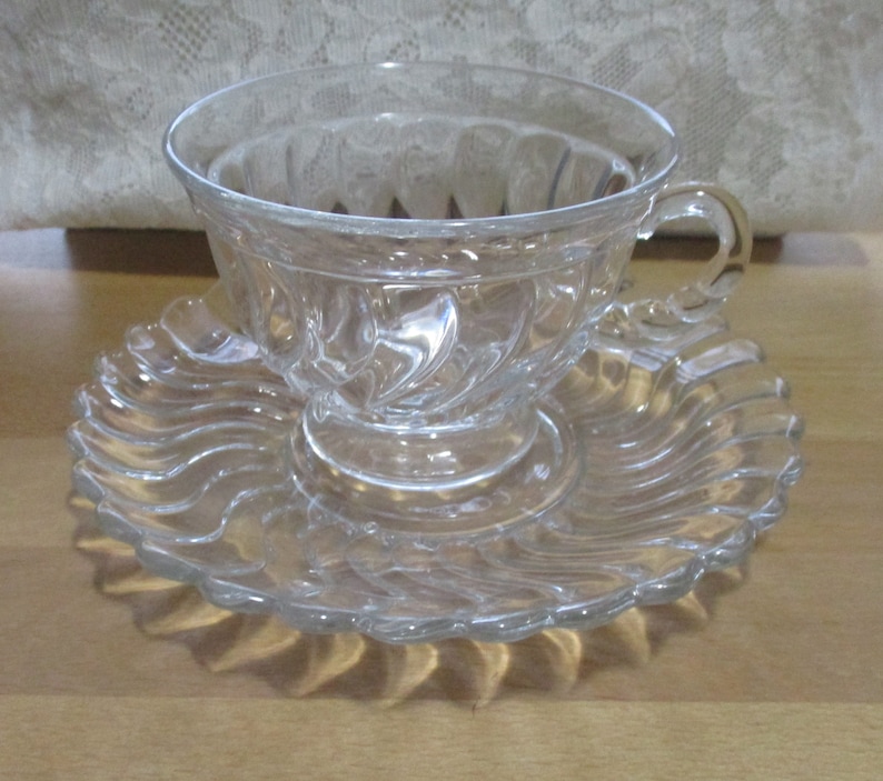 May include: A clear glass teacup and saucer set with a swirl design.