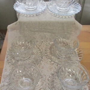 May include: Set of six clear glass teacups and saucers with a swirl design. The cups and saucers are arranged on a white lace tablecloth.