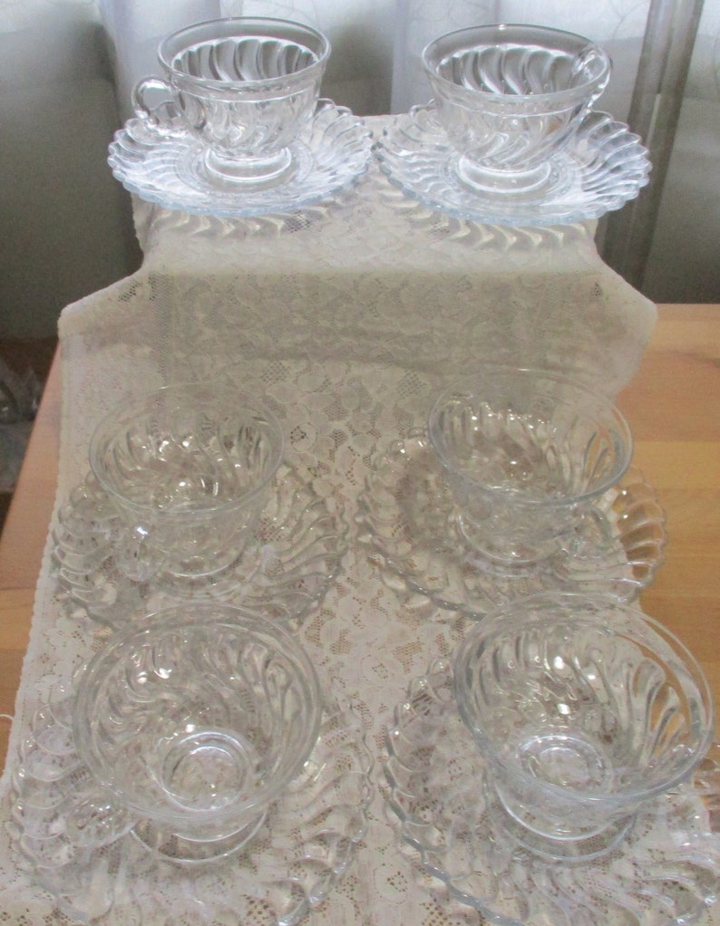 May include: Set of six clear glass teacups and saucers with a swirl design. The cups and saucers are arranged on a white lace tablecloth.