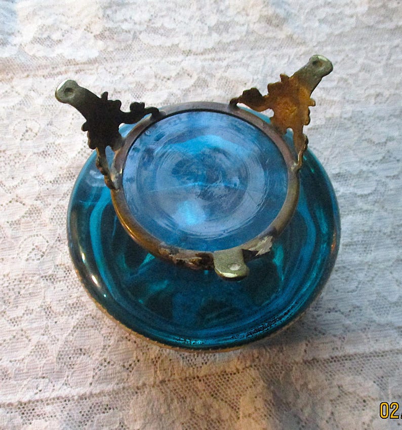 Op de afbeelding: Een vintage turquoise glazen kom met een messing rand en decoratieve details. De kom heeft een ronde vorm en een wervelend patroon in het glas. De messing rand heeft sierlijke details, wat bijdraagt aan de antieke uitstraling.
