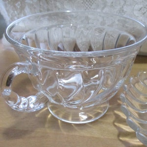 May include: A clear glass teacup with a fluted design and a handle. The cup is sitting on a wooden surface.
