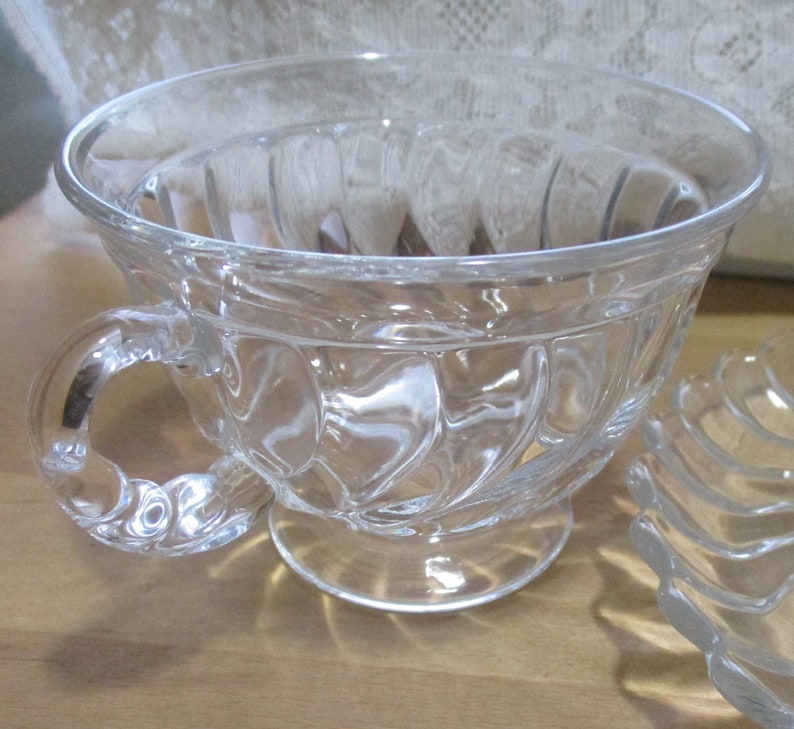 May include: A clear glass teacup with a fluted design and a handle. The cup is sitting on a wooden surface.