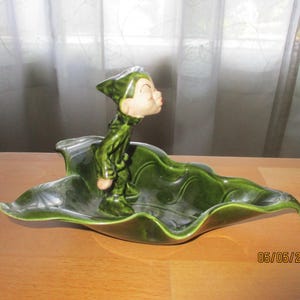 May include: A vintage ceramic figurine of a green elf standing on a large, leaf-shaped dish. The elf has a tan face and is wearing a green outfit and hat. The dish is a deep green color and has a glossy finish. The figurine is on a wooden table.