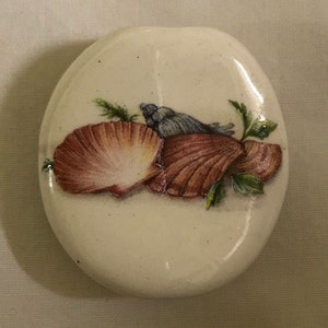 May include: A white ceramic oval dish with a painted design of three seashells and green seaweed. The seashells are brown, pink, and blue.