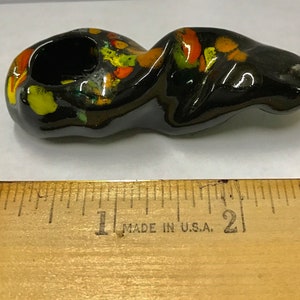 Może przedstawiać: Czarna ceramiczna fajka z kolorową glazurą. Fajka ma około 5 cm długości i okrągły ustnik. Glazura jest mieszanką żółtego, pomarańczowego i zielonego.
