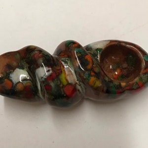 Może przedstawiać: Brązowa ceramiczna fajka z wzorem spiralnym i kolorowymi plamkami. Fajka ma czarkę na jednym końcu i długą, wąską rurkę na drugim.