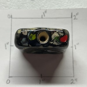 May include: A rectangular, ceramic bottle stopper with a dark base and colorful accents. The stopper is approximately 1 inch wide and 2 inches long. The design includes red, green, blue, and white spots. A central hole is visible.