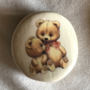 以下が含まれることがあります： 2匹の茶色のぬいぐるみクマが描かれた白いセラミックボタン。大きなクマはピンクのリボンをしています。