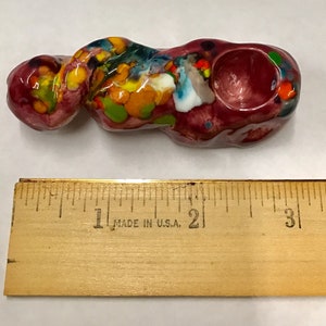 Może przedstawiać: Mała, kolorowa, ceramiczna fajka z czerwoną podstawą i misą. Fajka ma około 10 cm długości i jest w różnych kolorach, w tym żółtym, zielonym, niebieskim i białym.