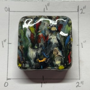 May include: A square, colorful glass art piece, approximately 1 inch by 1 inch. The artwork features a mix of colors including green, blue, red, and yellow, with a design that resembles a landscape or abstract scene.