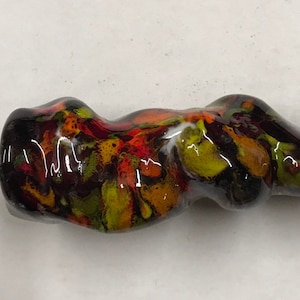 Może przedstawiać: Błyszcząca, abstrakcyjna rzeźba o płynnej, organicznej formie. Element charakteryzuje się mieszanką kolorów, w tym czarnym, czerwonym, pomarańczowym i żółtym, tworząc efekt marmuru. Rzeźba jest prawdopodobnie ceramiczna lub szklana.