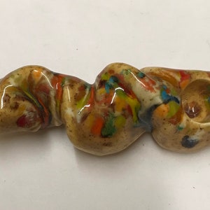 Może przedstawiać: Ceramiczna fajka z brązową, białą, pomarańczową i niebieską glazurą w kropki. Fajka ma kształt długiej, cienkiej, skręconej rurki z kominkiem na jednym końcu.