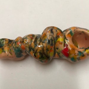 Może przedstawiać: Ręcznie malowana ceramiczna fajka z wirującym, abstrakcyjnym wzorem w odcieniach brązu, zieleni, żółci, pomarańczy i czerwieni. Fajka ma czarkę na jednym końcu i ustnik na drugim.